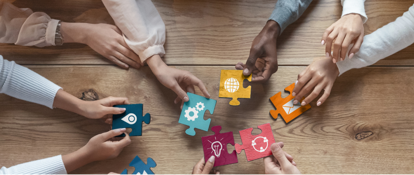 A group of hands working together to assemble a colorful puzzle with each puzzle piece featuring a different icon, symbolizing collaboration and teamwork